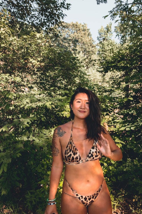 Tattooed Asian female in bikini standing in floral grove and looking at camera while enjoying sunny day