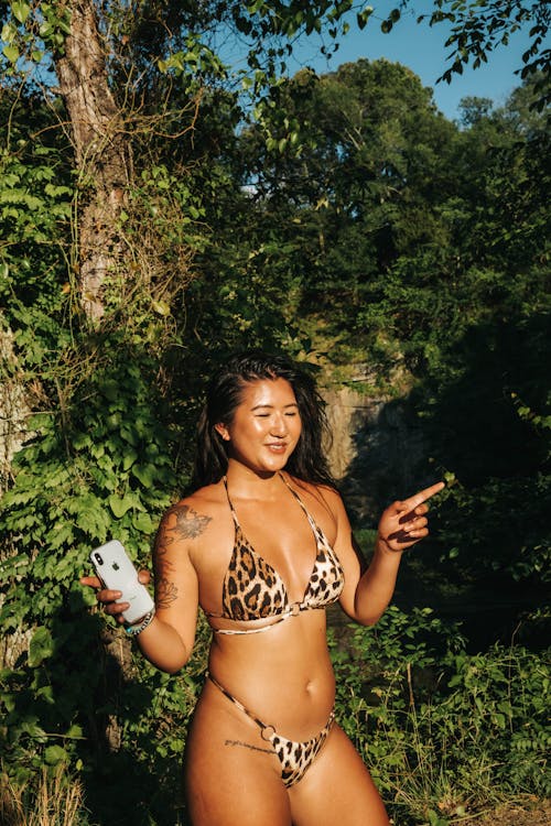 Free Joyful Asian female wearing swimsuit in leopard print standing with smartphone in green garden and dancing Stock Photo