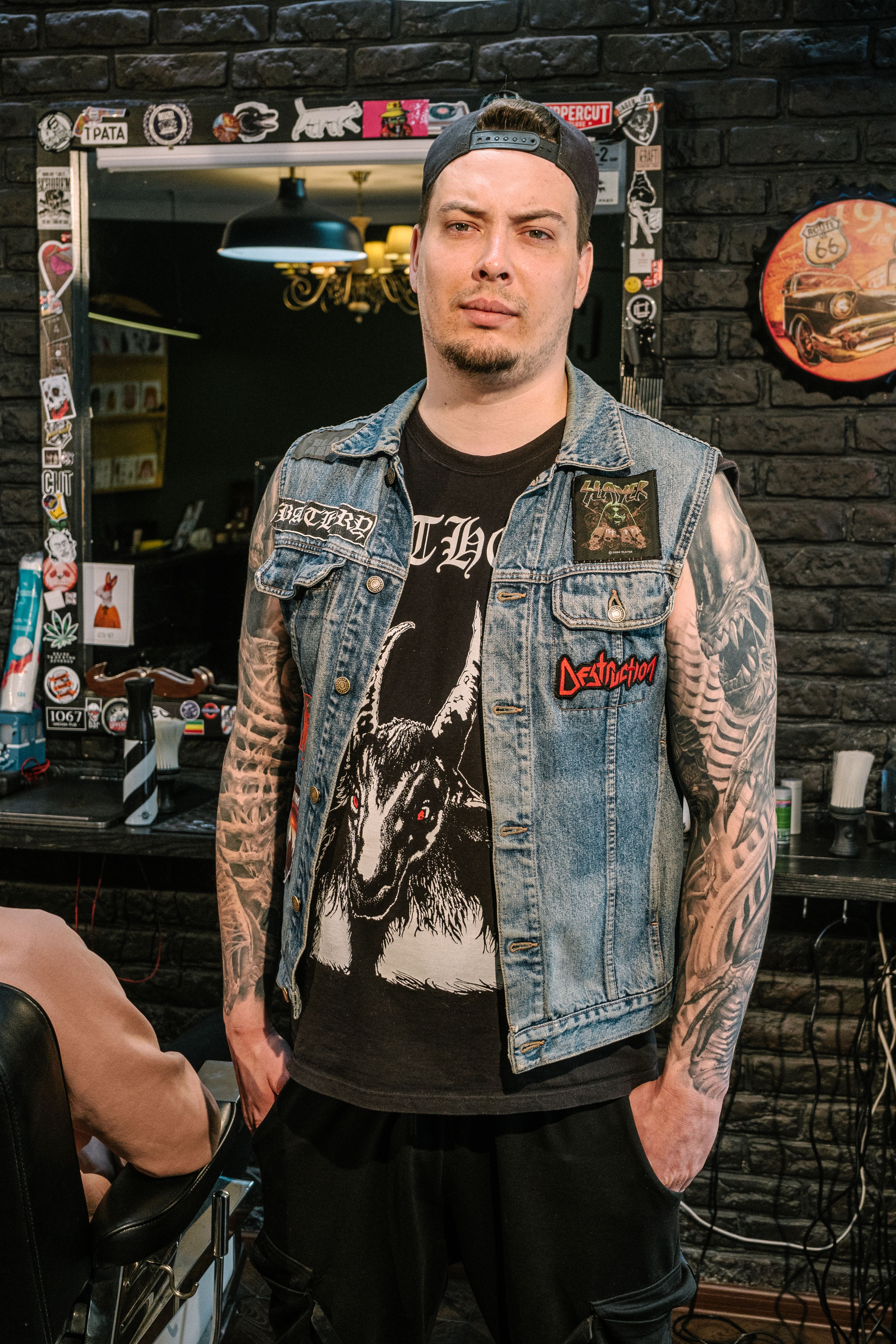 Man with tattoos in denim vest posing confidently in a trendy barbershop setting.
