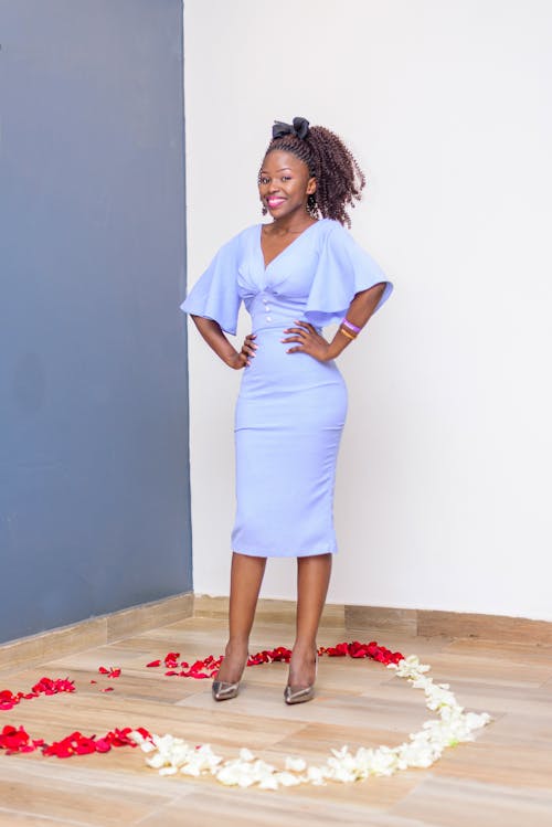 Full body stylish African American female in classy blue dress standing with hands on waist on floor in heart made with rose petals and looking at camera with toothy smile