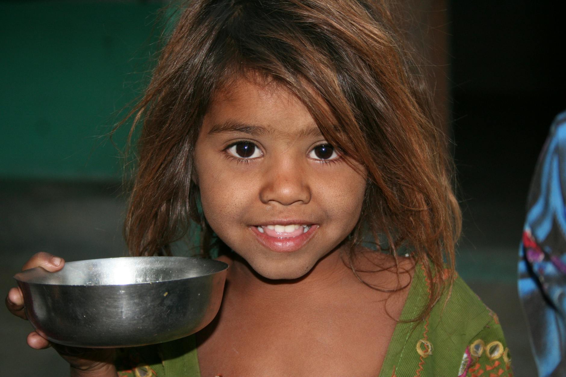 A young girl smiling while holding a metal bowl indoors, reflecting joy and innocence.
