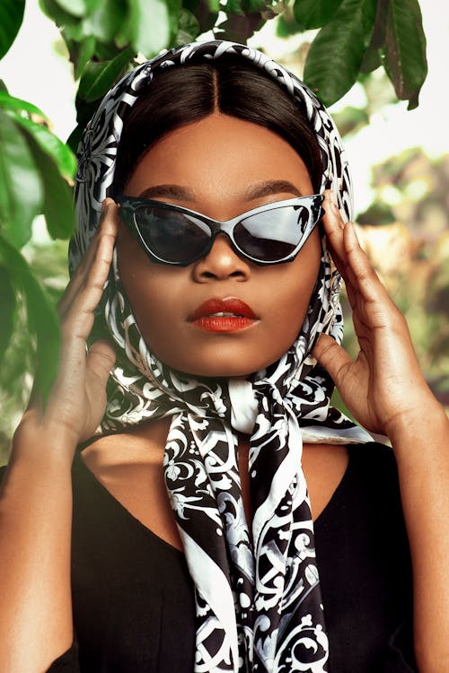 Fashionable African American female in stylish silk headscarf standing near lush tree and touching trendy sunglasses temples