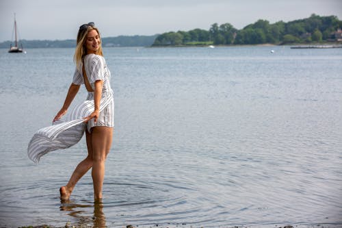 Side view full length content slim female in stylish maxi summer dress standing barefoot in shallow sea against picturesque landscape