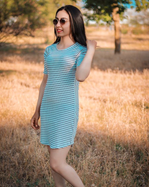 Free Positive young female in striped dress and stylish sunglasses touching hair and standing on grassy lawn in sunny countryside Stock Photo