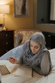A Muslim woman in a hijab lies on her bed, using a laptop for work or study in a cozy bedroom setting.
