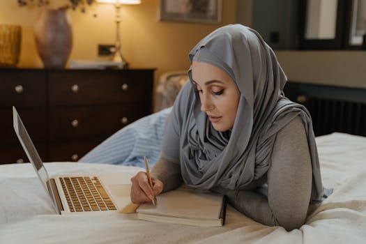 A Muslim woman in a hijab studying on her bed with a laptop and taking notes. Ideal for education and work-from-home visuals.