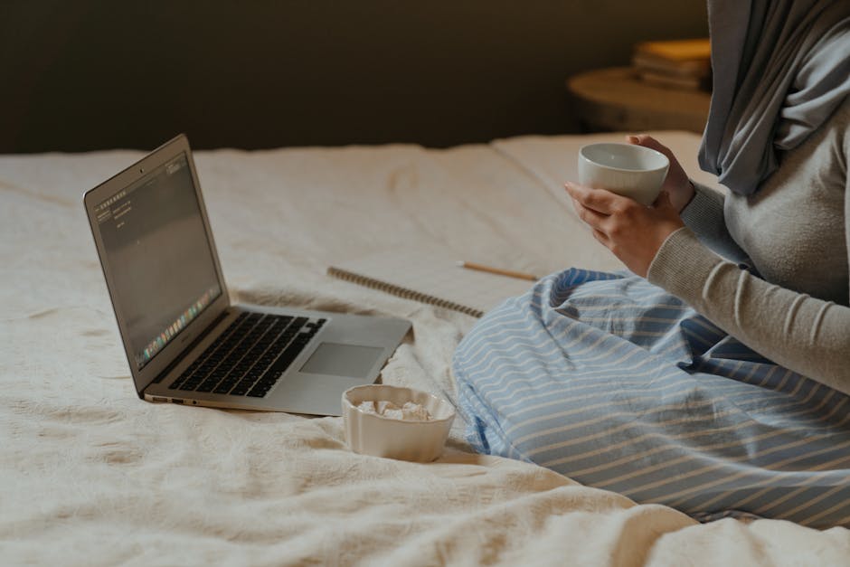 A woman working remotely from her bed with a laptop, tea, and notepad.