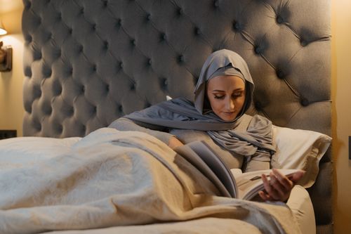 Free A woman in traditional Islamic attire reads a book while relaxing in bed. Stock Photo