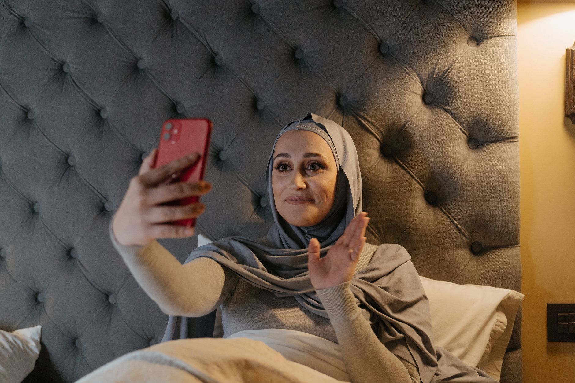Mujer con hiyab sentada en una cama, haciendo una videollamada con su smartphone rojo y saludando con la mano