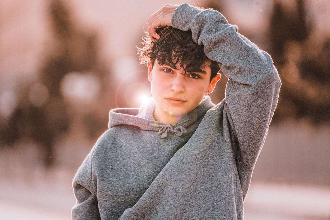 Attentive male teenager in trendy hoodie standing with raised arm while looking at camera in back lit