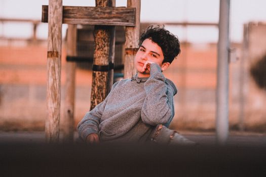 Contemplative male adolescent in trendy wear leaning on hand while sitting on bench in town and looking at camera