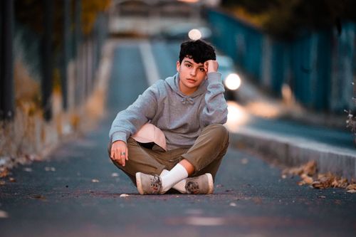Free Teen in trendy wear with cap sitting with crossed legs leaning on hand on roadway in town while looking at camera Stock Photo