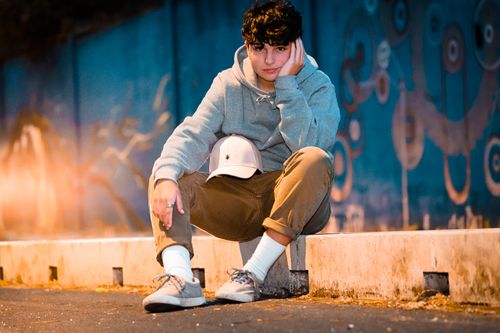 Free Ground level of crop stylish male adolescent in contemporary wear with cap sitting on barrier leaning on hand and looking at camera in evening Stock Photo