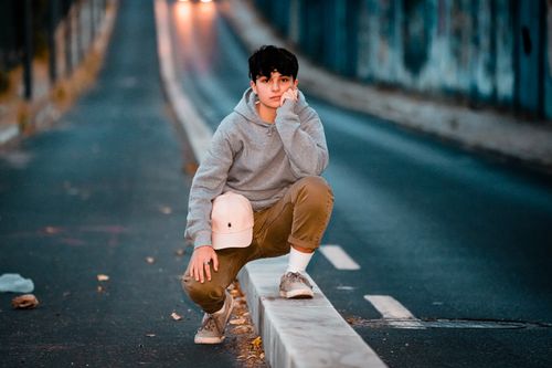 Free Contemplative male teen in trendy clothes with cap squatting on roadway barrier while leaning on hand and looking at camera in twilight Stock Photo