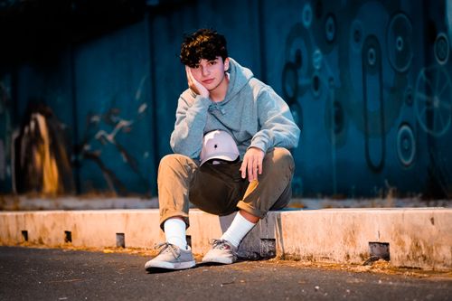 Free Ground level of young dreamy male teen in trendy wear sitting leaned on hand near city road and looking at camera Stock Photo