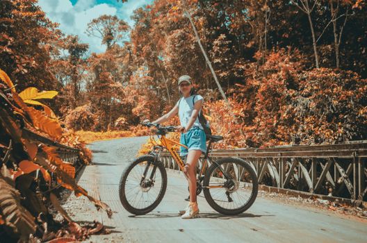 Cheerful female traveler with bicycle on roadway in earphones looking at camera between bright trees in fall
