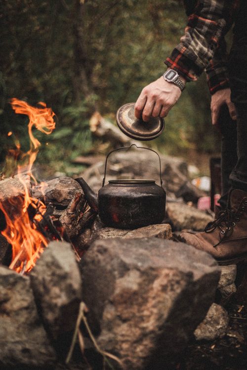 Free A hand placing a lid on a kettle over a vibrant campfire in an outdoor setting. Stock Photo