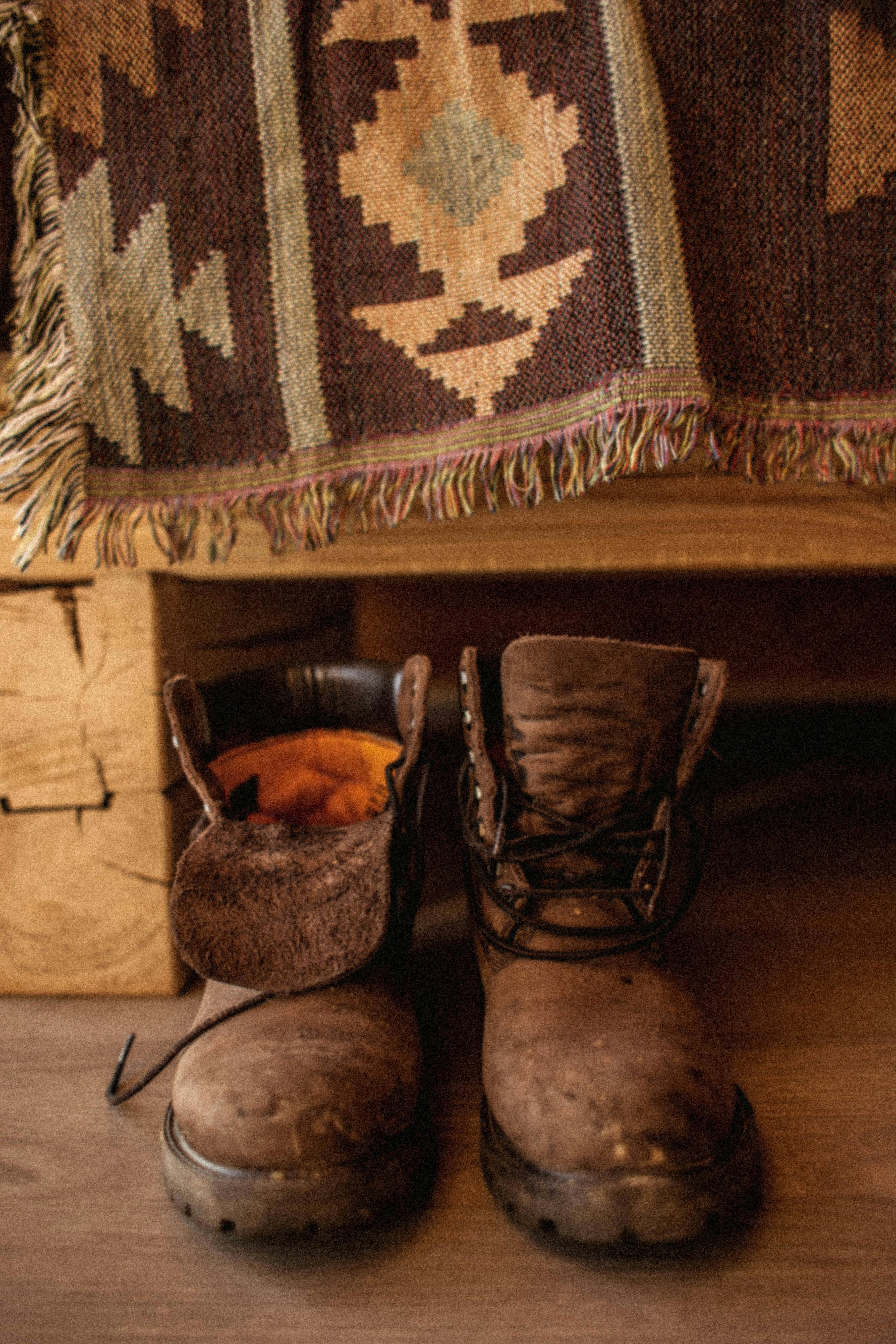 A warm and textured close-up of worn leather boots on a wooden floor with a patterned textile backdrop.