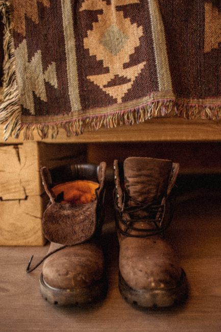 A warm and textured close-up of worn leather boots on a wooden floor with a patterned textile backdrop.