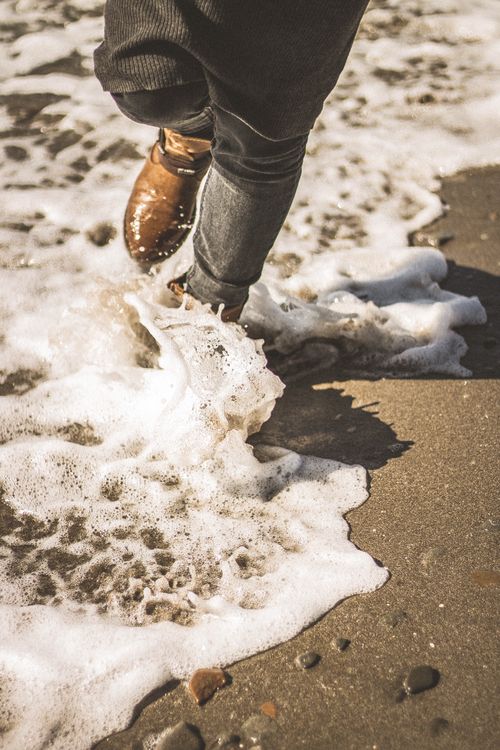 Free A person wearing jeans and boots walking in sea foam on a sandy beach. Stock Photo