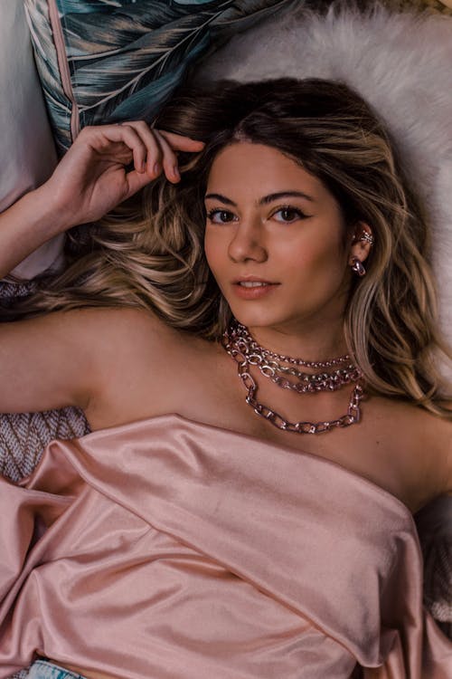 From above of young contemplative sensitive female in accessories touching hair while lying on bed at home and looking at camera