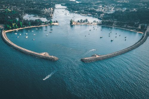 Free Drone view of breakwater and lagoon with motor boats on ocean behind trees and buildings in town Stock Photo