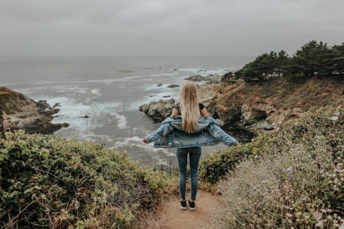 Free Back View Of Woman in Blue Denim Jacket and Blue Denim Jeans Standing On A Cliff Stock Photo