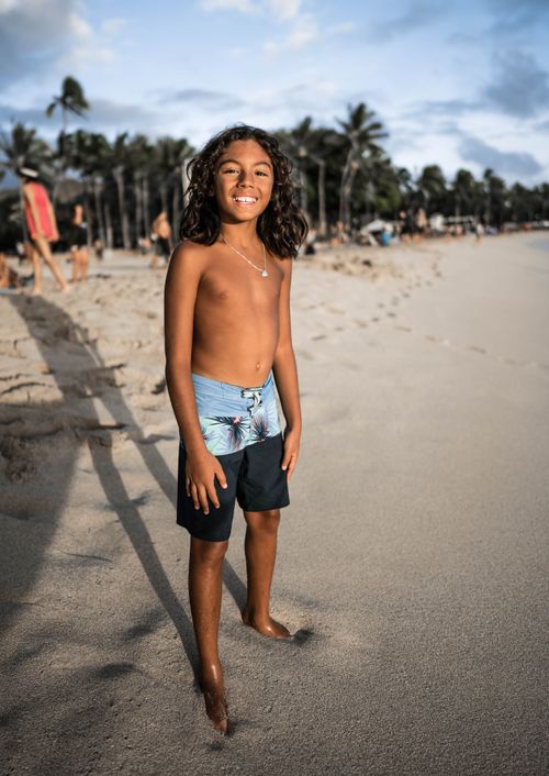 Free Full body cheerful Hispanic boy with long curly hair in swimming shorts standing on tropical sandy beach Stock Photo