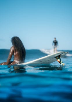 Back view anonymous African American female resting on surfboard on azure seawater near blurred surfer