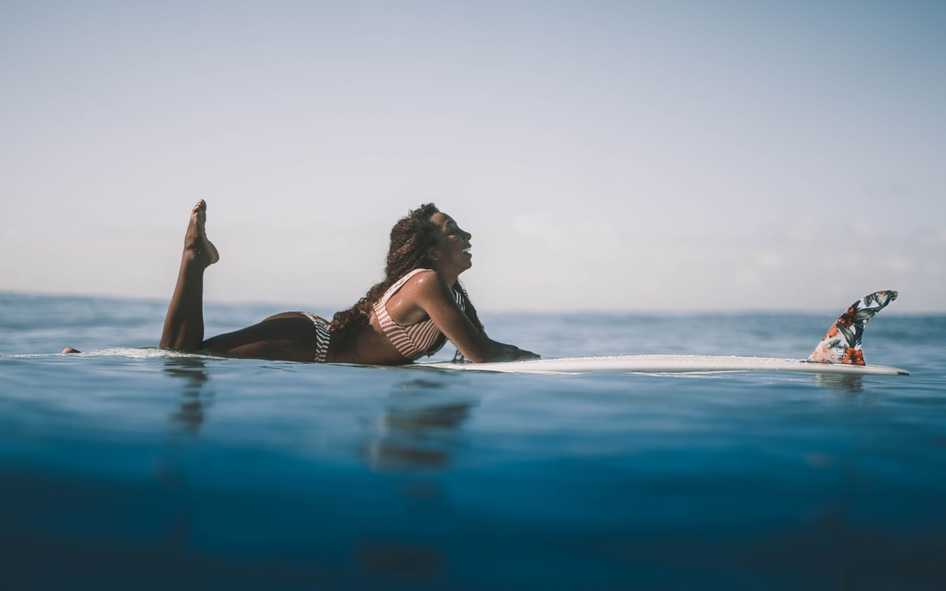 Side view smiling African American female in stylish swimwear lying gracefully on surfboard on azure seawater under clear sky