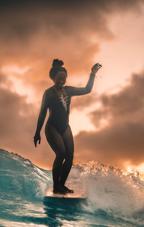 Free Full body of black female traveler in swimwear standing on surfboard while floating in wavy ocean against sunset sky Stock Photo