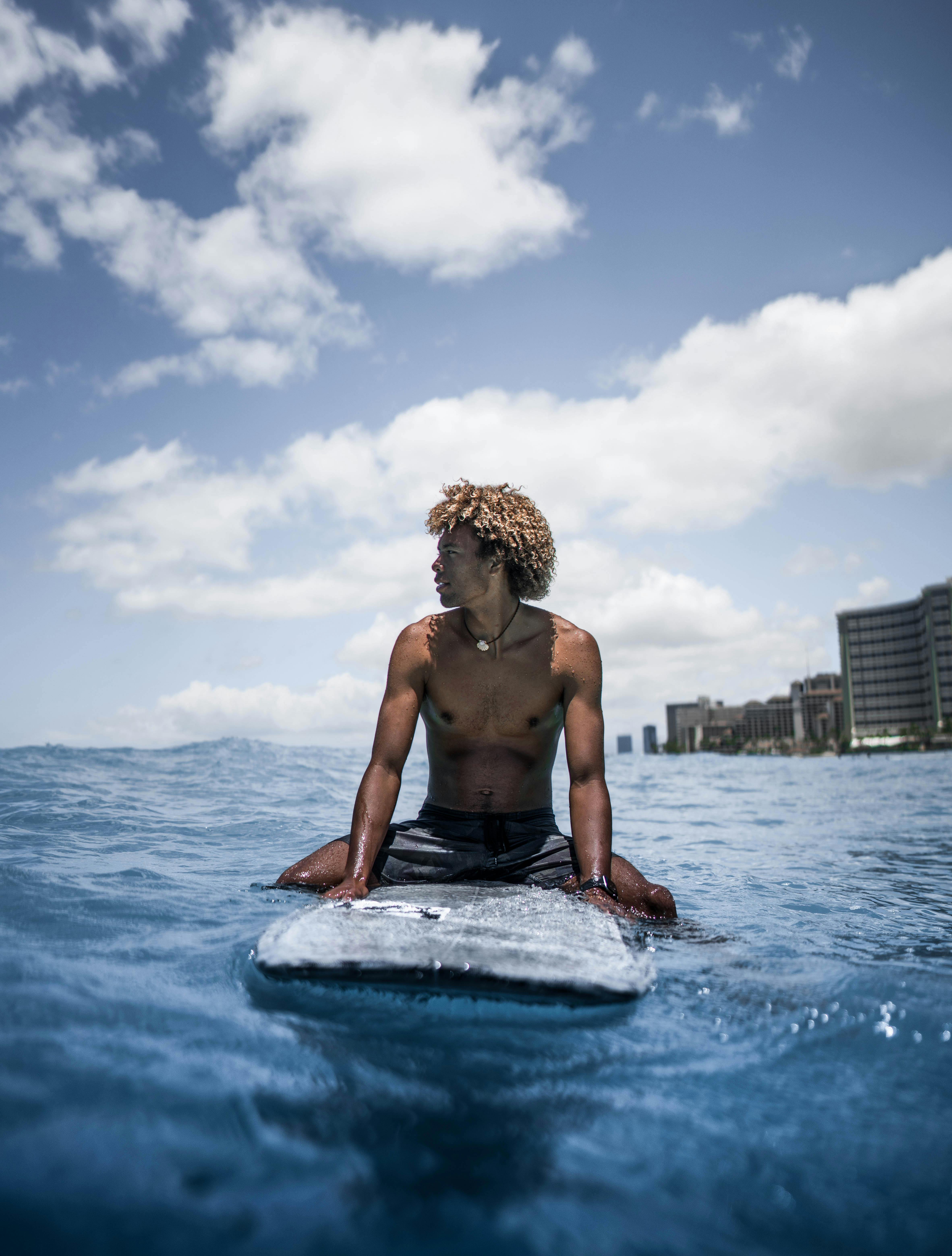 一名赤裸上身、留着非洲发型的年轻黑人男性运动员坐在冲浪板上，置身于波涛汹涌的大海中，目光移向别处