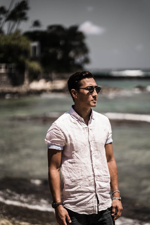 Young handsome ethnic male tourist in casual clothes and sunglasses standing on ocean beach and looking away