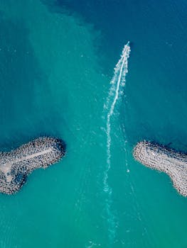 Aerial shot capturing a boat sailing through turquoise waters between two breakwaters.