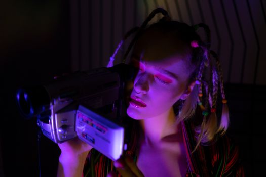 A fashionable woman with braids using a vintage camcorder under vibrant lighting.