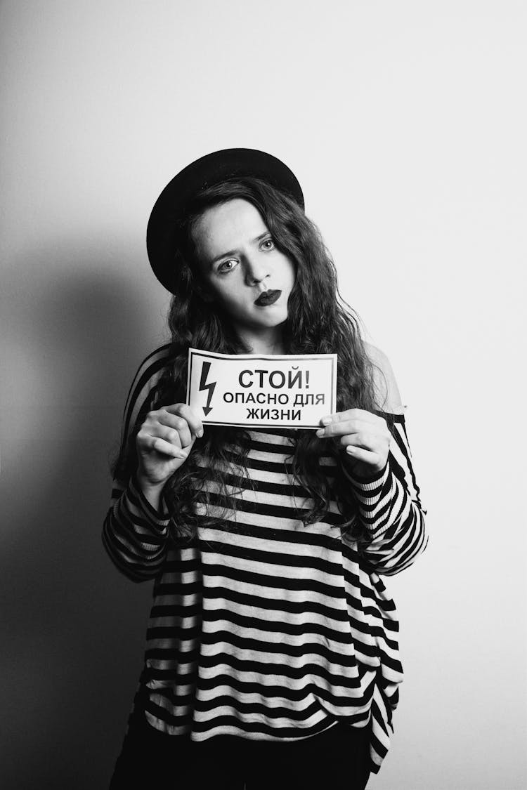 A Woman In Striped Long Sleeve Holding A Sign While Seriously Looking At Camera