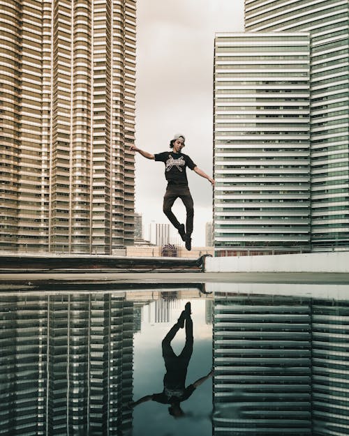 Free Full body of young ethnic male in casual outfit jumping on street between modern skyscrapers against cloudy sky Stock Photo