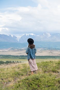 A woman in a denim jacket and hat stands on grassy hillside, overlooking a mountain landscape.