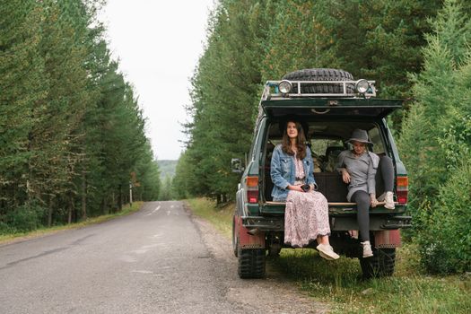 Two women sit on the back of an SUV parked by a forest road, enjoying nature.