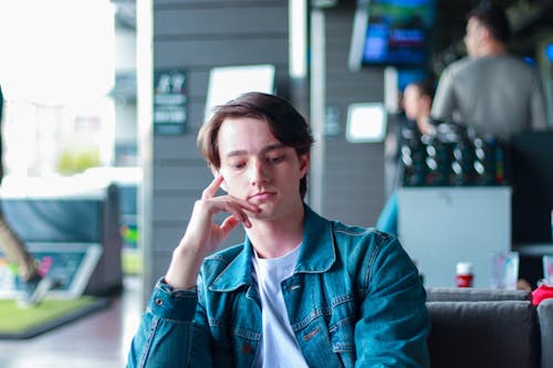 Thoughtful young male employee in casual wear touching face and looking down while sitting on comfortable couch in modern cafe during break
