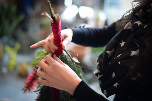 High angle of crop anonymous female designer wrapping red ribbon on floral bouquet  while creating decoration for event in workshop