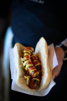 High angle of crop anonymous person with hot dog with sausages drizzled with ketchup and mustard in soft bun