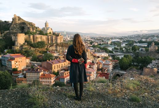 Woman overlooks Tbilisi cityscape with historic fortress and churches under a pastel sky.