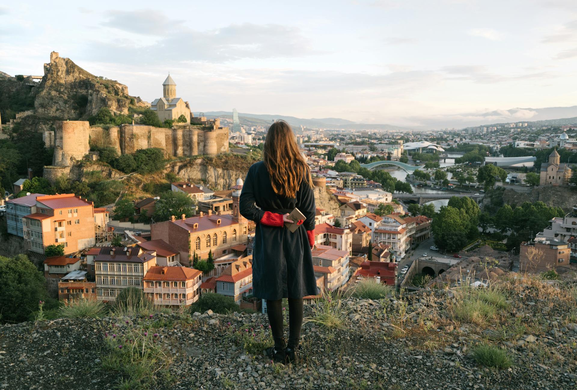 Woman overlooks Tbilisi cityscape with historic fortress and churches under a pastel sky.