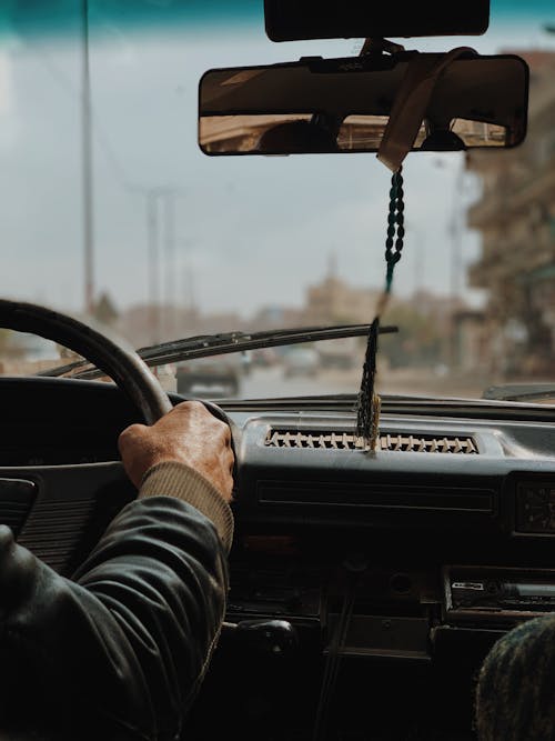 Crop unrecognizable male driving automobile with decorative beads on mirror on roadway in town