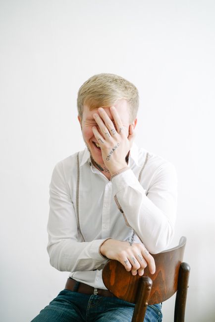 A man laughing with a tattooed hand covering his face, sitting on a wooden chair.