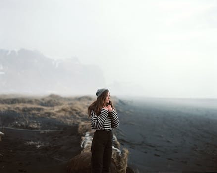 A woman in a striped sweater and beanie stands thoughtfully on a misty, deserted beach.