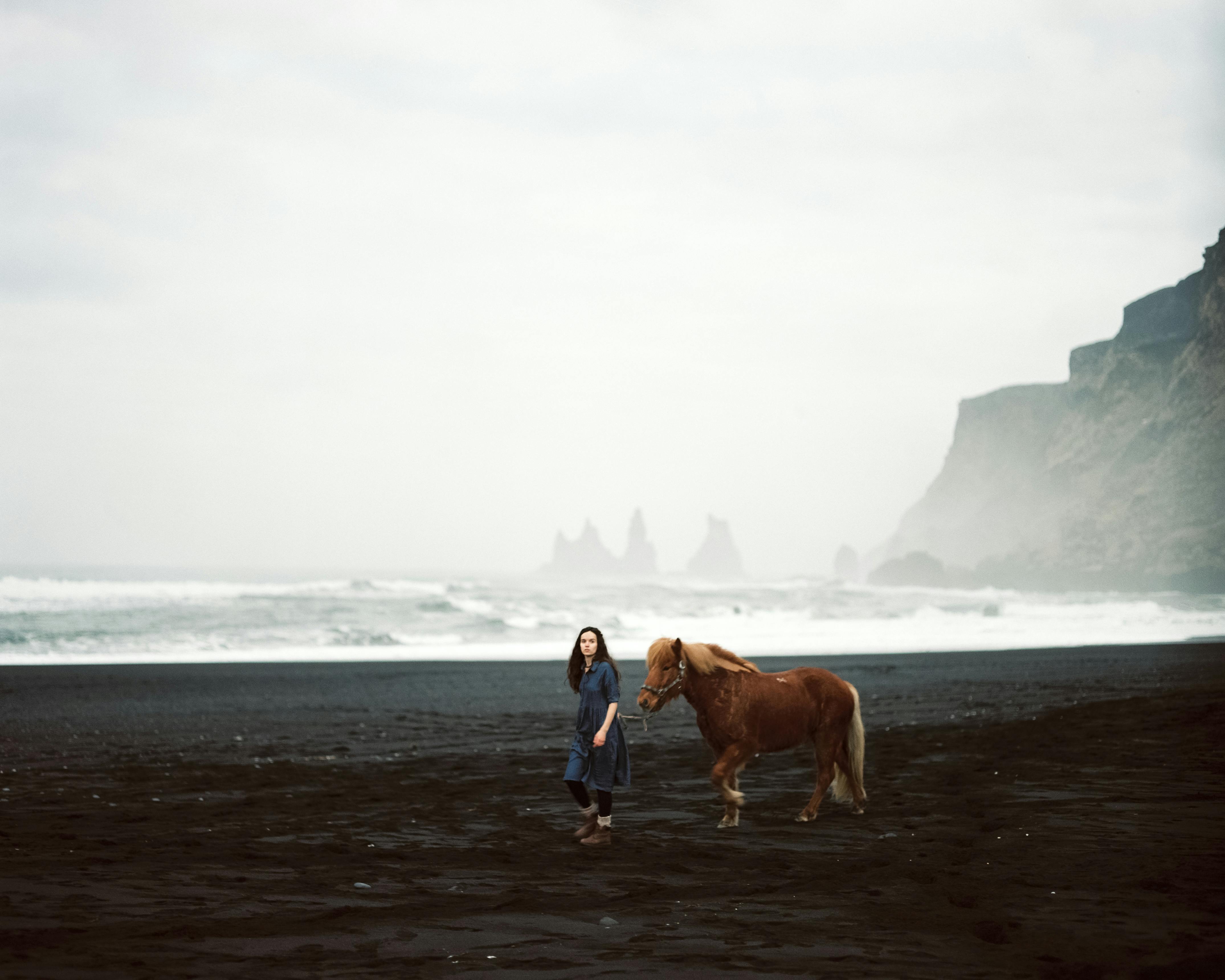 A woman leads a horse along a misty black sand beach with cliffs in the background.