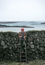 Woman in Red Knit Cap and Green Jacket Standing on Rock Formation Near Sea