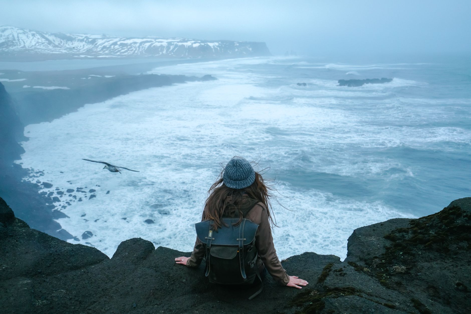 A woman sits on a cliff edge, overlooking the ocean with waves crashing below, creating a serene and adventurous scene.